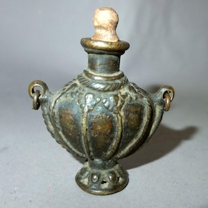 Peut inclure: Petite bouteille antique de couleur bronze avec un bouchon en liège. La bouteille a un corps renflé avec des détails décoratifs et deux petites poignées en anneau. La bouteille est probablement en métal et a un aspect vintage.