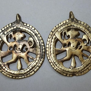 Op de afbeelding: Twee antieke goudkleurige hangers. Elke hanger heeft een centrale figuur in een cirkelvormig frame, omgeven door een kralenrand. De figuur lijkt een gestileerde menselijke of dierlijke vorm te zijn met uitgestrekte armen en benen. De hangers hebben een lus aan de bovenkant om op te hangen.