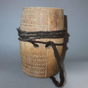 Peut inclure: Un baril en bois brun clair avec des motifs géométriques sculptés. Une épaisse corde noire est enroulée autour du milieu du baril et fixée par deux trous. Le baril a une forme légèrement conique.