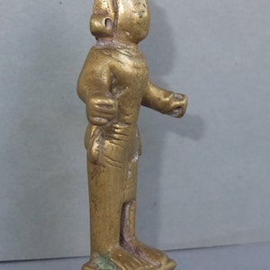 May include: A small, gold-toned metal figurine of a standing figure with a pointed headdress, extended arms, and a draped garment. The figure is mounted on a square, tiered base with visible etched detailing and a weathered, metallic finish.