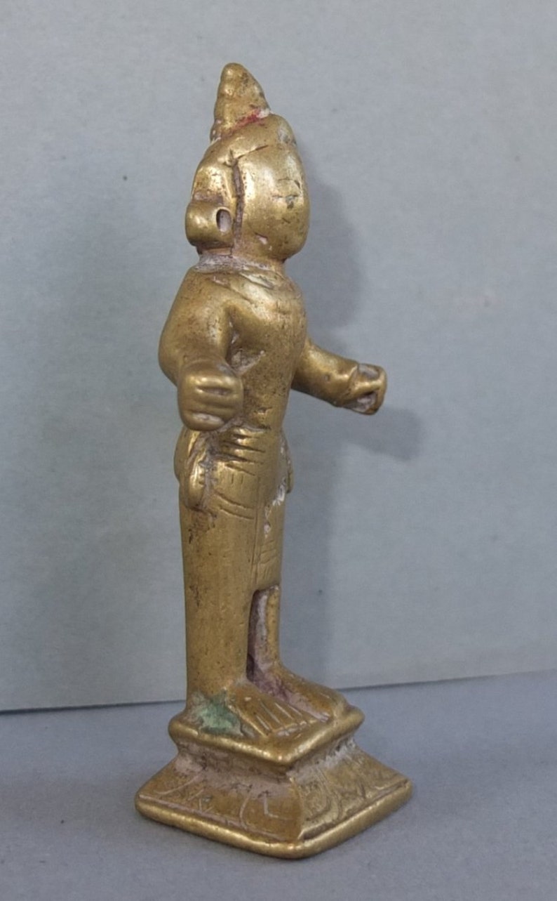 May include: A small, gold-toned metal figurine of a standing figure with a pointed headdress, extended arms, and a draped garment. The figure is mounted on a square, tiered base with visible etched detailing and a weathered, metallic finish.