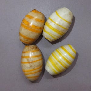 Può includere: Quattro perline ovali con base color crema e strisce orizzontali gialle e arancioni. Le perline hanno una finitura liscia e lucida e sono disposte su una superficie grigia. Queste perline potrebbero essere utilizzate per la creazione di gioielli o altri progetti artigianali.