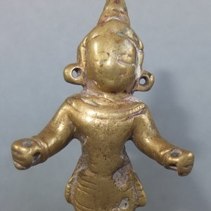 May include: A small, metallic gold-colored figurine with a smooth, rounded head, prominent ear ornaments, and outstretched arms. The figure features etched detailing on the lower torso and shows signs of wear with visible surface texture.