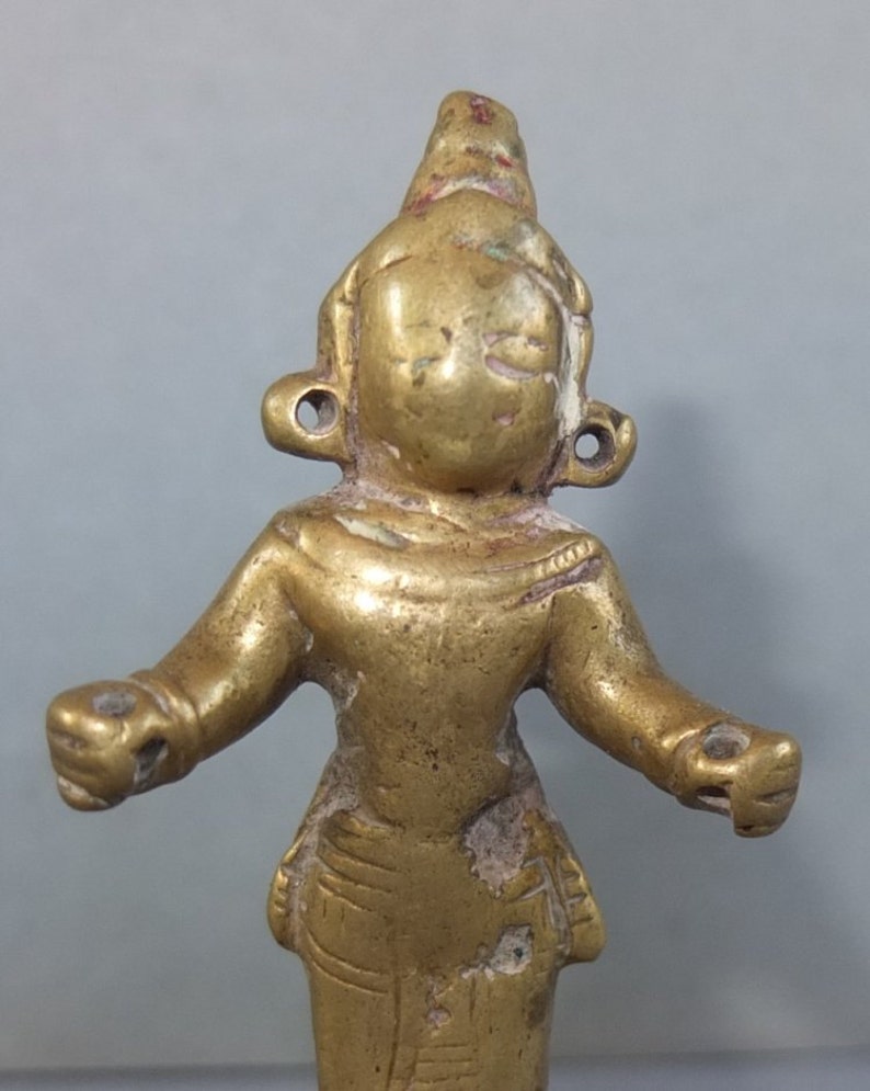 May include: A small, metallic gold-colored figurine with a smooth, rounded head, prominent ear ornaments, and outstretched arms. The figure features etched detailing on the lower torso and shows signs of wear with visible surface texture.