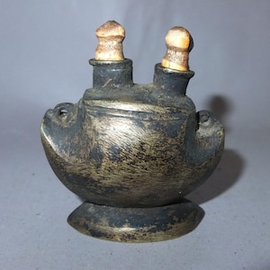 Peut inclure: Un récipient antique de couleur sombre avec deux becs, chacun surmonté d'un bouchon arrondi de couleur claire. Le récipient a un corps arrondi et une petite base circulaire. La surface a un aspect texturé et vieilli, suggérant qu'il s'agit d'un ancien artefact.