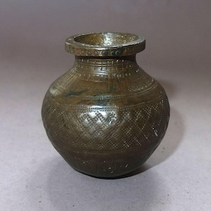Può includere: Un piccolo vaso antico color bronzo con corpo arrotondato e bordo leggermente svasato. La superficie è decorata con motivi incisi, tra cui motivi geometrici e floreali. Il vaso ha un aspetto invecchiato, che suggerisce età e utilizzo.