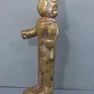 May include: A small, weathered gold-toned metal figurine standing on a square base. The figure features a pointed headdress, stylized facial features, and arms extended slightly outward. The surface shows signs of wear with visible texture.
