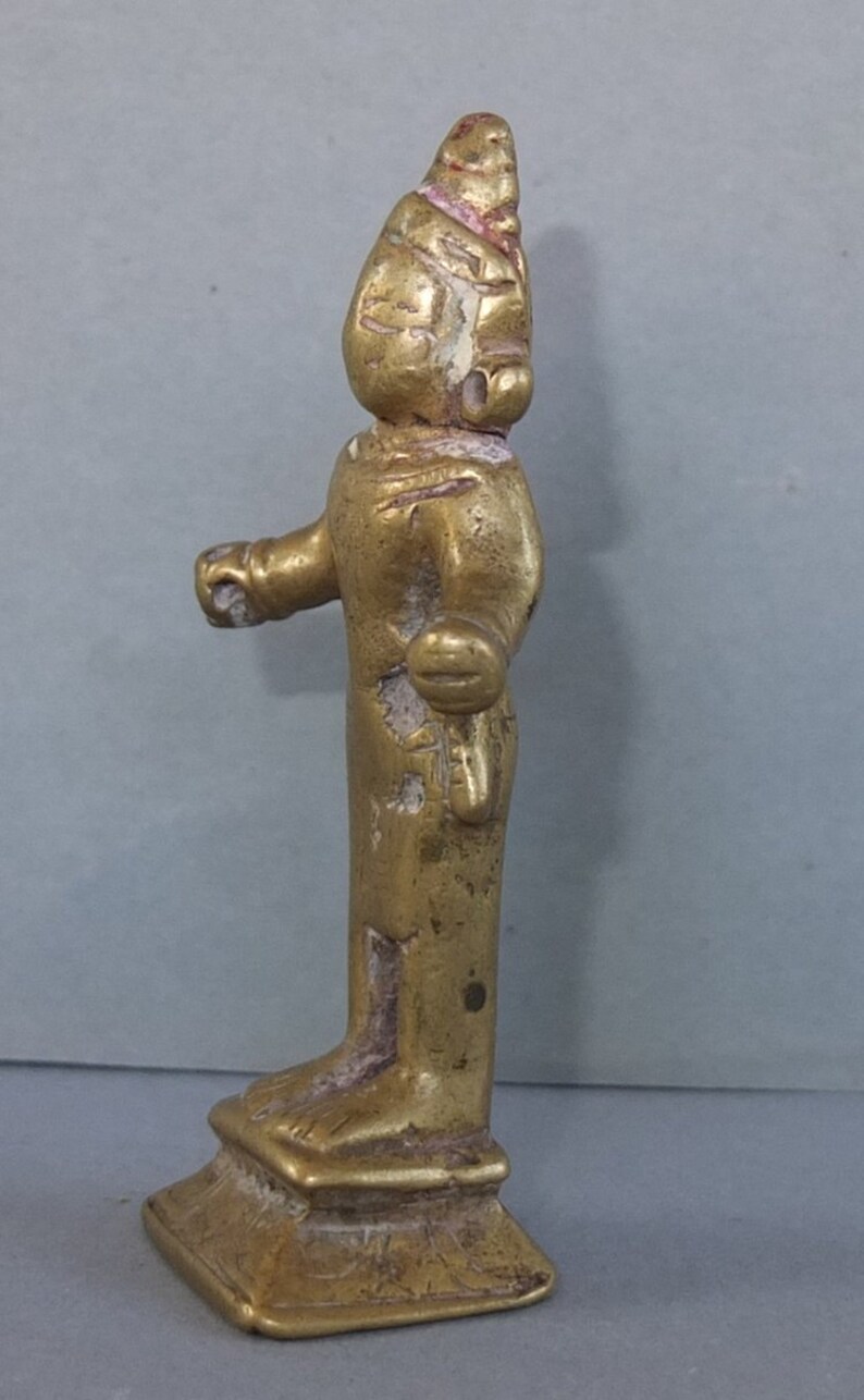 May include: A small, weathered gold-toned metal figurine standing on a square base. The figure features a pointed headdress, stylized facial features, and arms extended slightly outward. The surface shows signs of wear with visible texture.