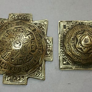 Peut inclure: Deux pièces décoratives antiques de couleur laiton. L'une est en forme de croix, l'autre carrée. Les deux présentent des motifs circulaires complexes avec des inscriptions et des éléments en forme d'étoile, suggérant une signification historique ou culturelle.