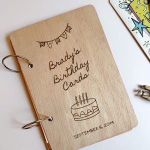 May include: A light-colored wooden scrapbook cover with two metal binder rings on the left side. The cover features a laser-engraved bunting banner, a birthday cake with four candles, and the text: Brady's Birthday Cards, SEPTEMBER 8, 2014.