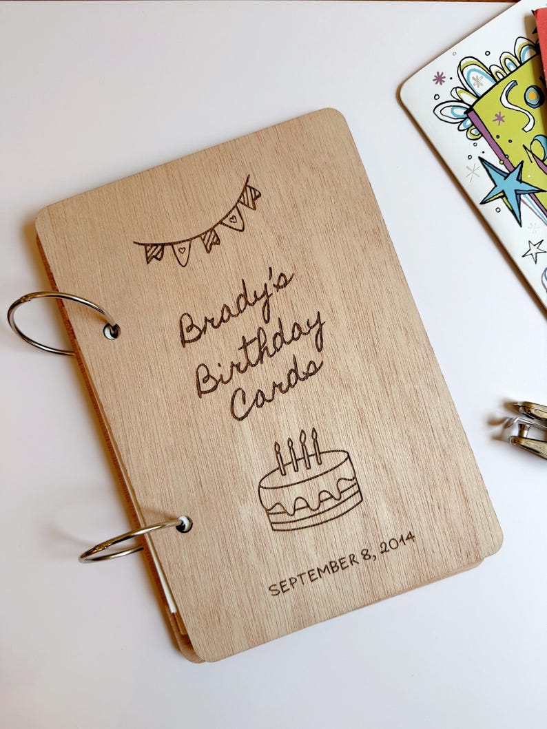 May include: A light-colored wooden scrapbook cover with two metal binder rings on the left side. The cover features a laser-engraved bunting banner, a birthday cake with four candles, and the text: Brady's Birthday Cards, SEPTEMBER 8, 2014.