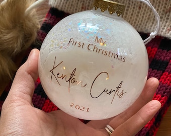 May include: A clear Christmas ornament with the text "My First Christmas" and the name "Kenton Curtis" in gold script, along with the year "2021". The ornament is filled with white glitter and held in a hand.