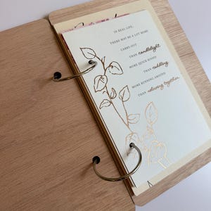 May include: A wooden-bound book with metal ring binding containing cream-colored pages. The visible page features a gold-foiled line drawing of a plant and text that reads: In real life, there may be a lot more carry-out than candlelight, more quick kisses than cuddling, more running around than relaxing together.