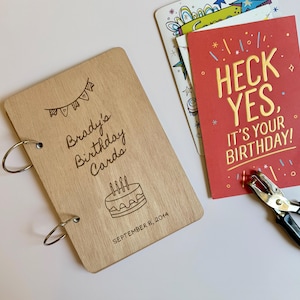 May include: A light-colored wooden cover for a card album with two metal binder rings, engraved with the text "Brady's Birthday Cards", a cake illustration, and "SEPTEMBER 8, 2014". Next to it is a red card with "HECK YES, IT'S YOUR BIRTHDAY!" text.