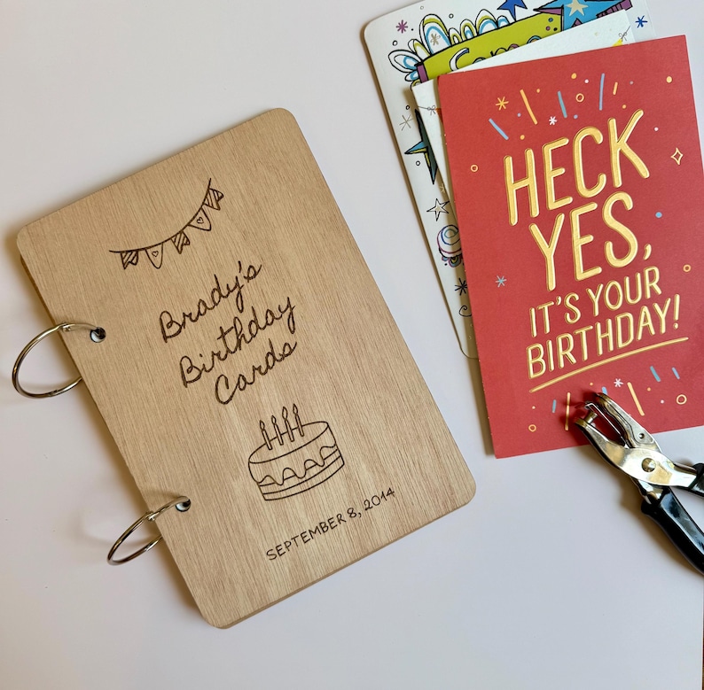 May include: A light-colored wooden cover for a card album with two metal binder rings, engraved with the text "Brady's Birthday Cards", a cake illustration, and "SEPTEMBER 8, 2014". Next to it is a red card with "HECK YES, IT'S YOUR BIRTHDAY!" text.