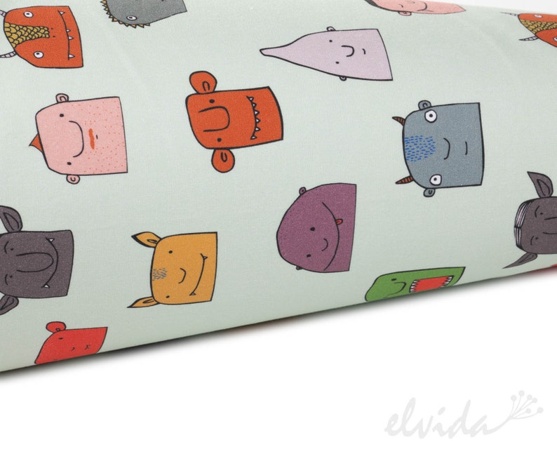 May include: A light green fabric with a repeating pattern of colorful cartoon monster faces. The monsters have different expressions and features, including horns, teeth, and eyes.