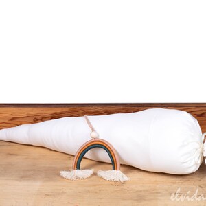 May include: A white fabric cone with a drawstring closure, lying on a wooden surface. A rainbow-colored yarn hanging decoration with tassels is in front of the cone.