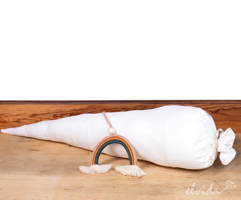 May include: A white fabric cone with a drawstring closure, lying on a wooden surface. A rainbow-colored yarn hanging decoration with tassels is in front of the cone.