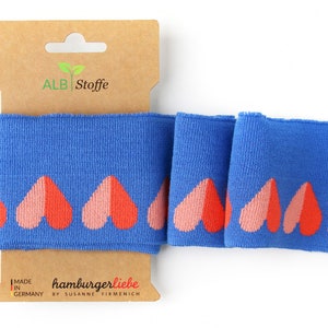 Könnte beinhalten: Blauer Strickstoff mit einem sich wiederholenden Muster aus rosa und orangen Herzen. Der Stoff ist mit "hamburgerliebe by susanne firmenich" und "made in germany" beschriftet.