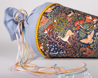 May include: A blue and yellow fabric cone with a forest scene print. The cone has a yellow pom-pom trim and a blue ribbon with a gold glitter pattern. The ribbon is tied in a bow and has long streamers in pink, blue, and yellow.