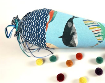 May include: A blue and white fabric cone with a whale and other sea creatures printed on it. The cone is decorated with a blue ribbon and has a blue and white wavy pattern at the top. There are colourful pom-poms scattered around the cone.