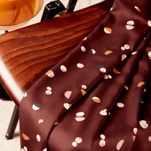 Könnte beinhalten: Ein burgunderfarbener Stoff mit einem abstrakten Muster in Weiß, Rosa und Gelb. Der Stoff ist gefaltet und über eine Holzoberfläche gelegt.