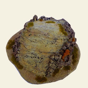 Puede incluir: Un modelo de terreno rocoso en miniatura con una parte superior plana, perfecto para juegos de mesa. El modelo está hecho de materiales marrones, grises y verdes, con una superficie texturizada.