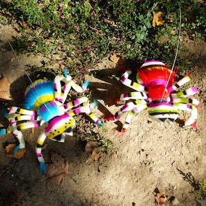 May include: Two colorful, handmade spider decorations with striped legs and bodies. One spider is blue, yellow, and white, while the other is red, pink, and white. They are set on a bed of dirt and leaves.