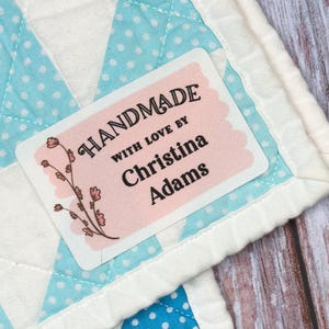May include: Rectangular fabric labels with floral illustrations and the text "HANDMADE WITH LOVE BY Christina Adams" or "MADE BY Christina Adams". One label is attached to a light blue and white polka dot quilt, while others sit in a pink heart-shaped dish.