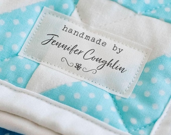 Op de afbeelding: Close-up van een handgemaakte quilt met een witte rand en een blauw-wit stippenpatroon. De quilt heeft een label met de tekst "handmade by Jennifer Coughlin". De tekst "FRAY PROOF BACKING NO NEED TO TURN UNDER EDGES" staat bovenaan.