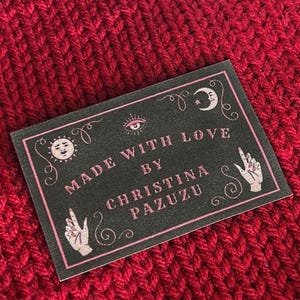 May include: A knitted fabric piece featuring a black upper section and a deep red lower section. A rectangular black label is attached, displaying the text "MADE WITH LOVE BY CHRISTINA PAZUZU" in pink, surrounded by illustrations of hands, a sun, and a moon.