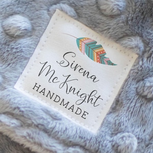 May include: A light gray dimpled minky fabric blanket featuring a white square label stitched to the corner. The label displays a colorful feather graphic and the text Sirena McKnight HANDMADE. The blanket has a soft, textured surface.
