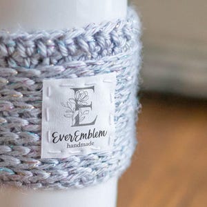 May include: A white ceramic travel mug featuring a light blue crocheted sleeve with subtle iridescent fibers. A white fabric label is stitched to the front, displaying the letter E, a floral illustration, and the text EverEmblem handmade.