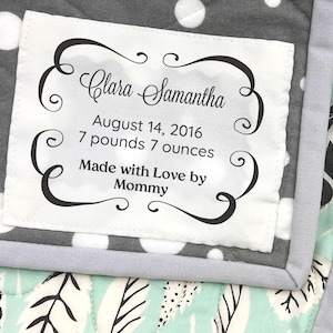 May include: A quilted fabric item with unfinished edges and a single layer of cotton. A white rectangular label features the text Clara Samantha, August 14, 2016, 7 pounds 7 ounces, and Made with Love by Mommy. The fabric has grey polka dots and leaf patterns.