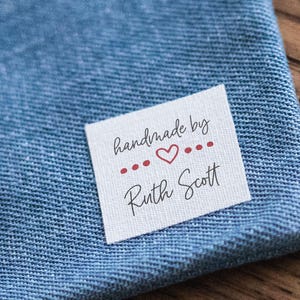 May include: A square white fabric clothing label featuring the black text handmade by Ruth Scott with a small red heart and dots in the centre, attached to a blue denim fabric. The label includes icons for no fray edges, machine wash and dry.