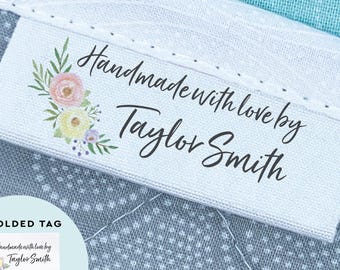 May include: A white fabric clothing label featuring a floral illustration in pink, yellow, and green, with the black text "Handmade with love by Taylor Smith". The label is folded and stitched onto a grey patterned fabric, with an inset showing the tag.