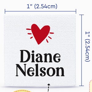 Könnte beinhalten: Ein quadratisches weißes Baumwolletikett mit den Maßen 2,54 cm mal 2,54 cm, das ein rotes Herzsymbol über dem schwarzen Text Diane Nelson zeigt. Ein kreisförmiger Einschub zeigt eine vergrößerte Ansicht des Drucks neben einem orangefarbenen Strick.