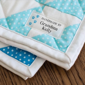 May include: A quilted fabric piece featuring a white and light blue polka dot pattern with a rectangular label sewn on top. The label displays the text HANDMADE BY Grandma Kelly with blue paw print icons. The top text reads FRAY PROOF BACKING.