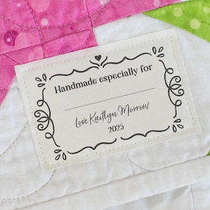 May include: A close-up of a handmade quilt label with the text "Handmade especially for" and "Love Kaitlyn Morrow 2025" in a decorative frame. The quilt features pink, white, and green fabric.