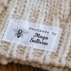 May include: A white rectangular fabric label sewn onto a beige knitted garment. The label features a black graphic of a bee on the left side and the black printed text "Handmade by Maya Sullivan" on the right side. The label has visible stitching.