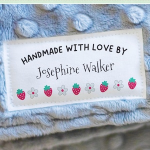 May include: A light blue, textured blanket with a white label that reads "HANDMADE WITH LOVE BY Josephine Walker." The label is decorated with strawberry and flower graphics. The text "Your Label, Your Way" is at the top, with a "Customize Now" button at the bottom.