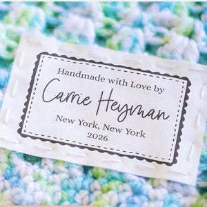 Peut inclure: Une étiquette blanche avec une bordure noire en pointillés, avec le texte "Handmade with Love by Carrie Heyman, New York, New York, 2026." L'étiquette est sur un fond crocheté bleu, vert et blanc. Les mots "Your Label, Your Way" sont en haut.