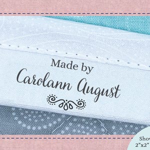 May include: A white fabric clothing label with black text reading Made by Carolann August and a decorative flourish. The label is shown folded on a blue patterned fabric. The set includes 20 labels, each measuring 2 inches by 2 inches before folding.