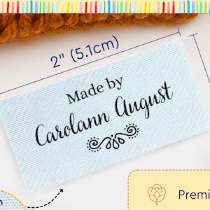 May include: A light blue rectangular cotton label with the black text Made by Carolann August and a decorative flourish. The label measures 2 inches by 1 inch. The image includes text stating Custom Cotton Labels for Handmade Creations.