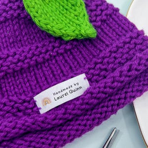 Può includere: Un berretto lavorato a maglia viola con un accento a foglia verde, dotato di un'etichetta in tessuto bianco con la scritta Handmade by Laurel Quinn e un'icona arcobaleno. Accanto, quattro etichette identiche su un piatto a forma di cuore e un uncinetto da 4,00 mm.