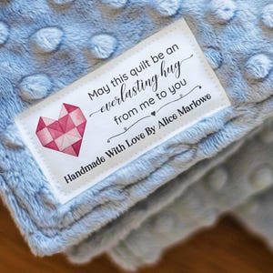 May include: A light blue minky blanket with a raised dot texture. A white rectangular fabric label is sewn onto the corner, featuring a pink geometric heart graphic and the text: May this quilt be an everlasting hug from me to you.