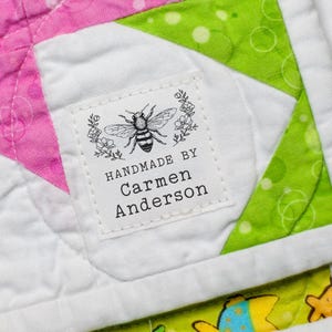 May include: A quilted fabric piece featuring a white, pink, and lime green geometric star pattern with a white fabric label sewn on. The label displays a bee illustration and the text HANDMADE BY Carmen Anderson in black ink.
