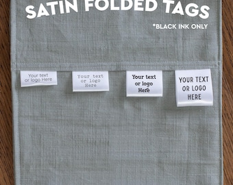 Könnte beinhalten: Vier gefaltete weiße Satin-Etiketten mit schwarzem Text auf einem salbeigrünen Stoffhintergrund. Die Etiketten zeigen den Text "Your text or logo Here" in verschiedenen Schriftarten. Das Bild enthält den weißen Text "SATIN FOLDED TAGS *BLACK INK ONLY".