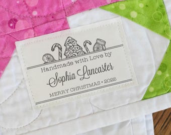 May include: A rectangular white fabric label stitched onto a quilted textile with pink and green patterns. The label features black printed text reading Handmade with Love by Sophia Lancaster MERRY CHRISTMAS 2025, with illustrations of a tree, candy canes, and a mitten.
