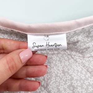 以下が含まれることがあります： Susan Heartsenという文字とHANDMADE WITH LOVEというフレーズ、そして小さなハートと針のアイコンが描かれた白い布製の衣類ラベル。ラベルは、グレーの花柄が施された白い生地の淡いピンク色の布製トリムに取り付けられています。